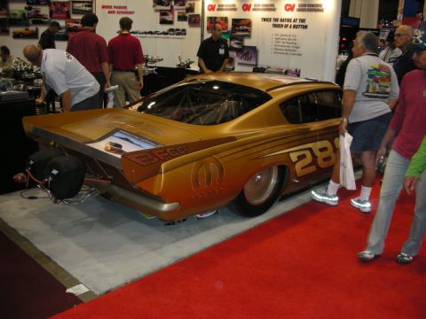 1998 Ford Crown Victoria - gold fastback drag-style car wearing number 28 and a Mopar logo, displayed on red carpet at an indoor trade show.