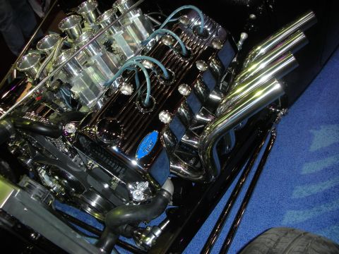1998 Ford Crown Victoria - chrome V8 with individual velocity stacks and polished lake-style headers mounted in a hot rod on blue carpet.