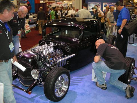 1998 Ford Crown Victoria - purple chopped highboy hot rod with an exposed chrome multi-carb V8, examined by spectators at an indoor show.