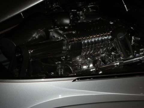 1998 Ford Crown Victoria - polished screw-type supercharger sits under the partly opened hood of a silver custom car in dim lighting.