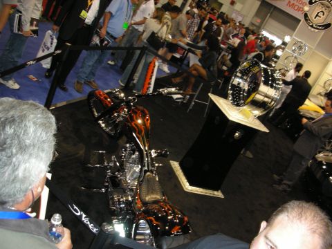 1998 Ford Crown Victoria - custom chopper motorcycle with orange flame paint sits on a black display platform at a crowded indoor show.