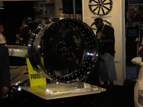 1998 Ford Crown Victoria - oversized chrome-lipped black bead-lock wheel displayed on a stand at a busy indoor automotive show booth.