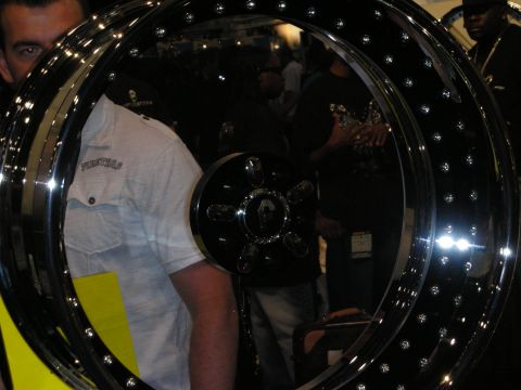 1998 Ford Crown Victoria - man in a white shirt and cap viewed through the center of a large chrome bead-lock display wheel at a show.