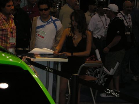 1998 Ford Crown Victoria - woman in a black dress signs at a booth table next to a green show car amid attendees under dim event lighting.