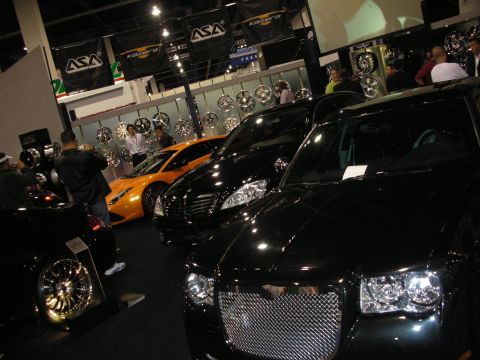 1998 Ford Crown Victoria - black Chrysler 300 with mesh grille beside an orange sports car at an indoor booth lined with display wheels.