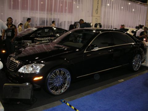 1998 Ford Crown Victoria - black Mercedes-Benz S-Class sedan on large chrome multi-spoke wheels displayed on blue carpet at an indoor show.