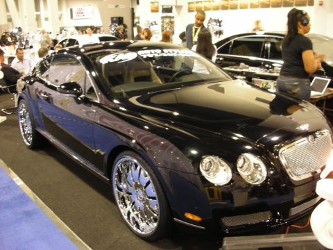 1998 Ford Crown Victoria - black Bentley Continental GT coupe on large chrome wheels with window decals on display at an indoor car show.