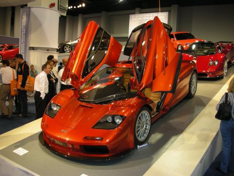 1998 Ford Crown Victoria - Orange sports car with butterfly doors open at an auto show, with other vehicles in the background.