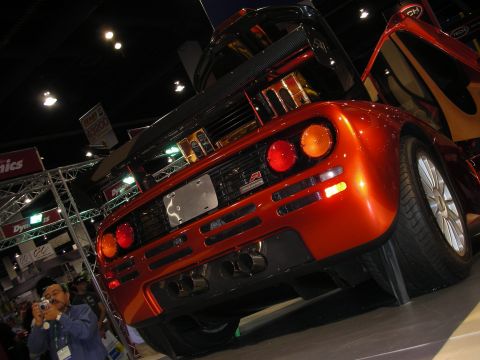 1998 Ford Crown Victoria - A bright orange McLaren F1 is displayed with its doors open, its rear design and quad exhaust pipes.