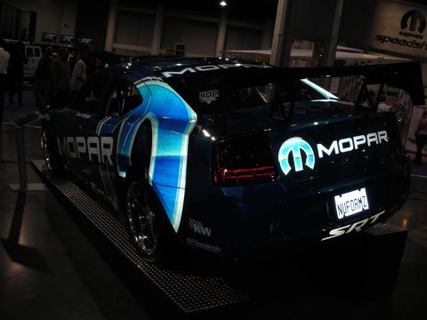 1998 Ford Crown Victoria - dark blue Dodge Charger SRT decked in Mopar graphics and a rear wing sits on a display platform at an indoor show.
