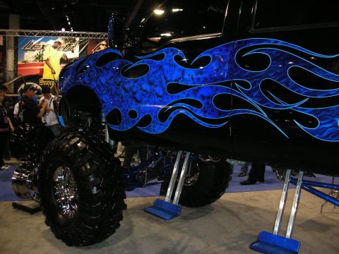 1998 Ford Crown Victoria - tall lifted truck with blue tribal-flame airbrush paint and large off-road tires sits on jack stands at an indoor show.