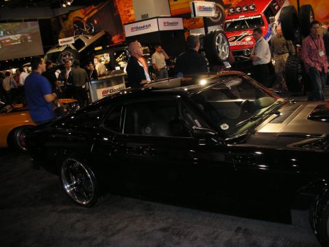 1998 Ford Crown Victoria - glossy black fastback muscle car on polished wheels sits among crowds and exhibitor booths at an indoor show.