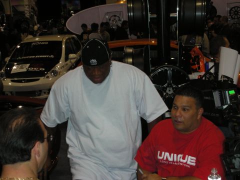 1998 Ford Crown Victoria - Two men, one in a white shirt and cap and one in a red UNIQUE shirt, talk at a booth amid show cars and display wheels.