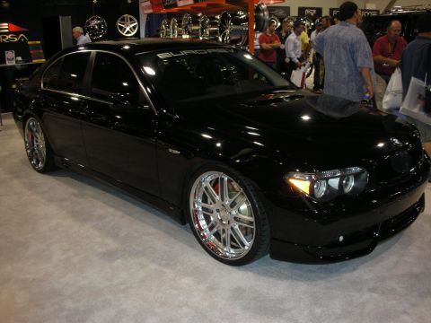 1998 Ford Crown Victoria - glossy black BMW 7 Series sedan lowered on polished multi-spoke wheels sits among show crowds and wheel displays.