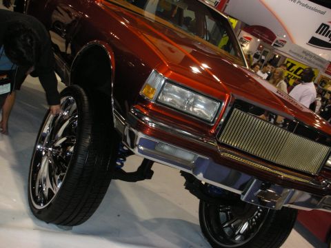1998 Ford Crown Victoria - candy-orange custom sedan on chrome wheels sits raised on coilover suspension with a gold grille at an indoor show.