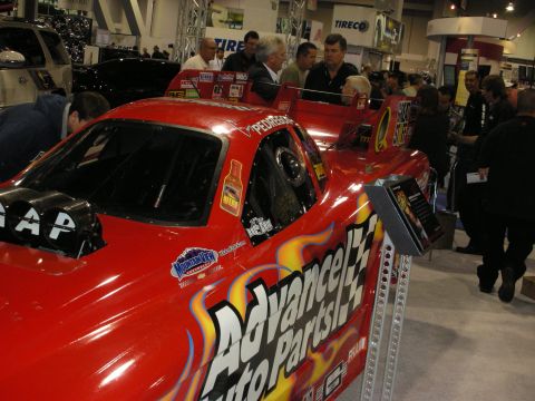 1998 Ford Crown Victoria - bright red Advance Auto Parts-liveried race car sits on display surrounded by people at a busy indoor automotive trade show.