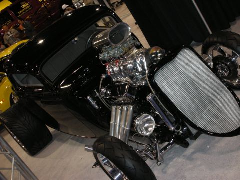 1998 Ford Crown Victoria - black hot rod displays a polished supercharged engine and exposed chrome front suspension on a show floor beside a yellow car.