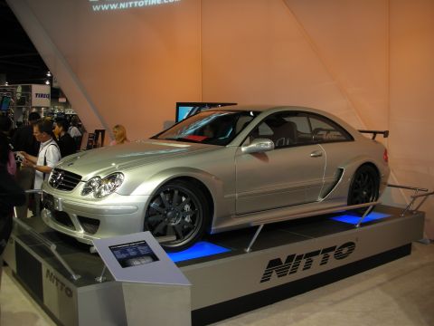 1998 Ford Crown Victoria - silver Mercedes-Benz CLK coupe with a rear wing sits raised on a blue-lit Nitto Tire display platform at an indoor show.