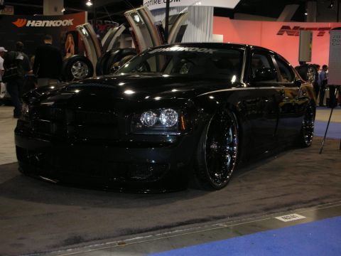 1998 Ford Crown Victoria - glossy black Dodge Charger sits low on dark multi-spoke wheels at a Hankook tire booth on an indoor show floor.