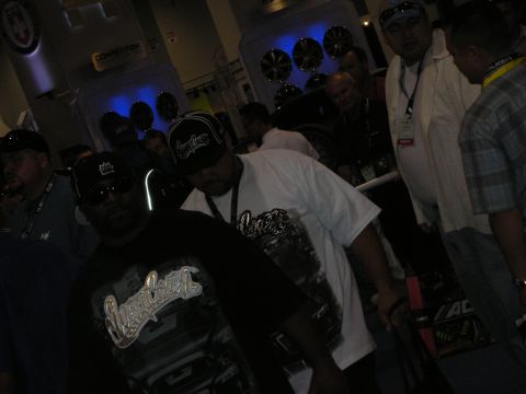1998 Ford Crown Victoria - dimly lit indoor crowd in caps and graphic T-shirts gathers near blue-lit audio speaker displays at a trade show booth.