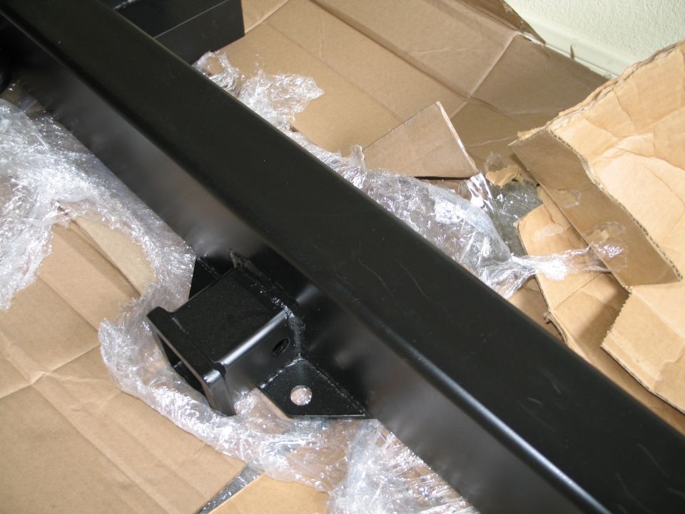 Black metal rear bumper with a hitch receiver, wrapped in plastic and resting on cardboard