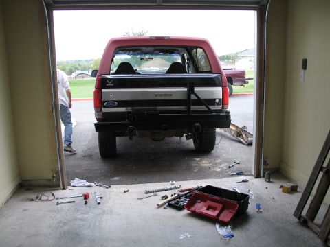 1995 Ford Bronco - Rear view of a 1995 Ford Bronco in a garage, showing a custom bumper and spare tire carrier. Another vehicle is visible outside.