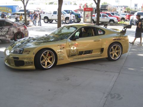 1998 Ford Crown Victoria - A gold sports car with numerous racing decals, including 'TOP SECRET', is parked outdoors with other vehicles in the background.