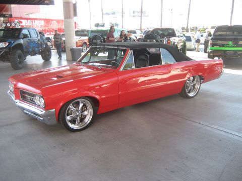 1998 Ford Crown Victoria - A classic red convertible is parked indoors with other vehicles and people around.