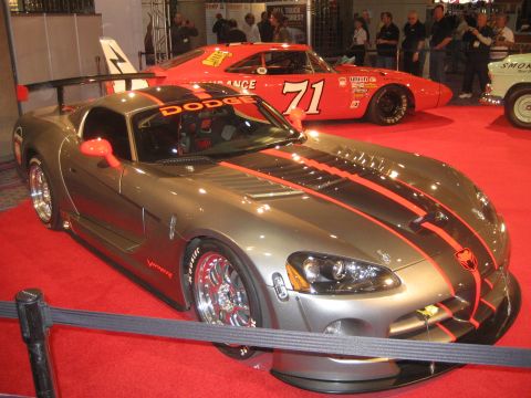 1998 Ford Crown Victoria - A gray Dodge Viper with prominent orange racing stripes and a rear spoiler is displayed on a red carpet, with a red race car visible behind…