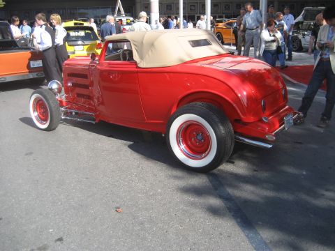 1998 Ford Crown Victoria - A bright red convertible hot rod with whitewall tires is displayed outdoors among other vehicles and onlookers.