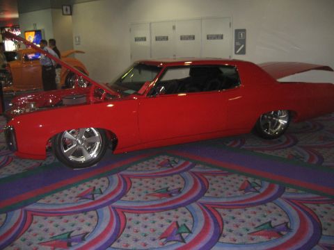 1998 Ford Crown Victoria - A red classic car is displayed indoors with its hood up, next to another orange vehicle.
