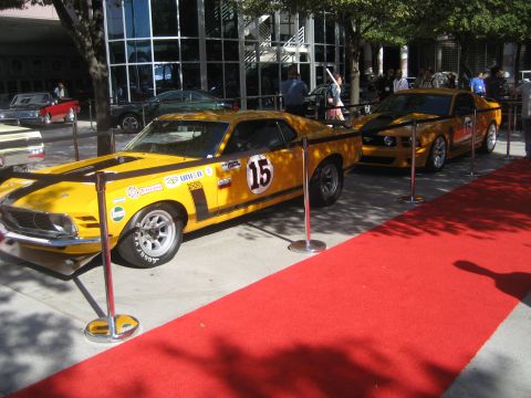1998 Ford Crown Victoria - Two vintage yellow race cars are displayed on a red carpet in front of a modern building.