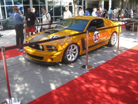 1998 Ford Crown Victoria - A yellow Ford Mustang with racing decals is displayed on a red carpet with stanchions.
