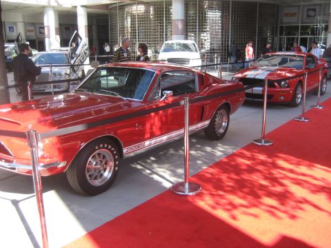 1998 Ford Crown Victoria - Two red Ford Mustangs, one a 1967 GT500, are displayed on a red carpet.