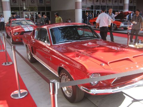 1998 Ford Crown Victoria - A red 1967 Shelby GT500 is displayed on a red carpet, with another red Mustang and an orange car in the background.