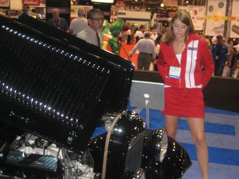 1998 Ford Crown Victoria - A woman in a red outfit stands next to a classic car with its hood open at an indoor event.