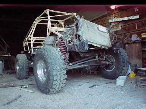 1984 Ford Bronco - Three-quarter front view of a bare tube-chassis buggy under construction in a wooden barn, with a fully welded roll cage, no body panels, e…