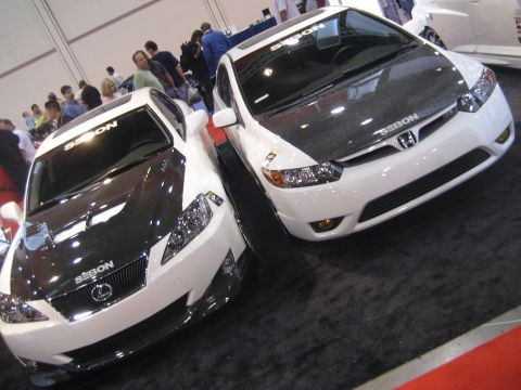 1998 Ford Crown Victoria - Two modified cars with carbon fiber hoods are displayed indoors on a black carpet.