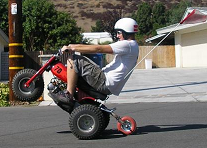 1900 Honda Big Red - A rider on a red three-wheeled ATV performs a wheelie on a paved road with houses in the background.