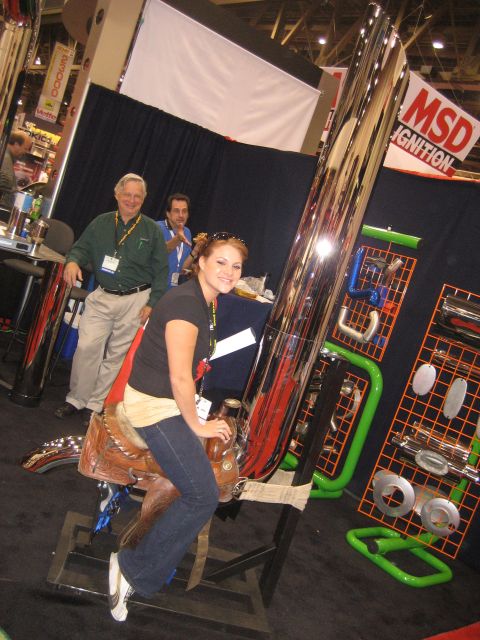 1998 Ford Crown Victoria - A woman poses on a saddle display at an indoor event, with a large chrome pipe and various automotive parts around her.