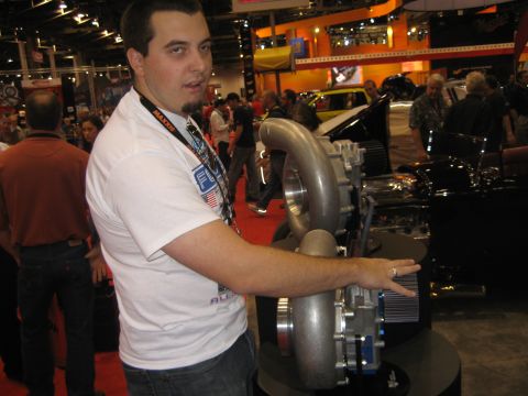 1998 Ford Crown Victoria - A man gestures towards a large supercharger and engine component display at an indoor event.