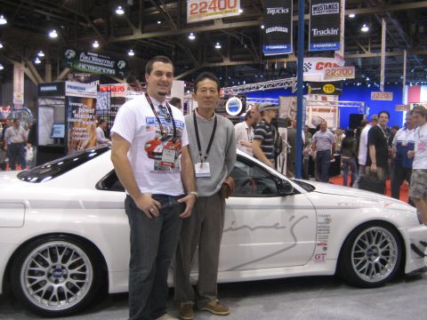 1998 Ford Crown Victoria - Two men stand beside a white sports car adorned with various racing decals and aftermarket wheels.