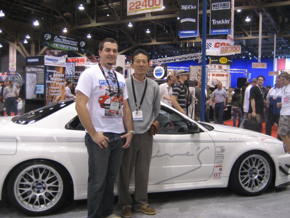 Two men posing beside a white Mine's sports car at a trade show