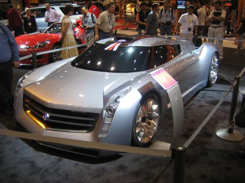 1998 Ford Crown Victoria - A futuristic silver concept car is displayed indoors, with people observing in the background.
