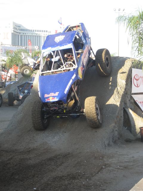 1998 Ford Crown Victoria - An off-road buggy navigates a challenging dirt obstacle course with spectators and buildings in the background.