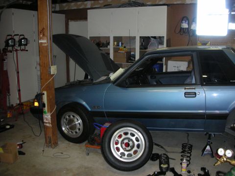 1990 Ford Mustang - A blue 1990 Ford Mustang coupe is shown in a garage with its hood open and suspension components laid out for service.