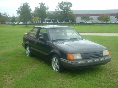 1989 Ford Escort - A dark 1989 Ford Escort coupe sits on a grassy lawn in front of a modern building.