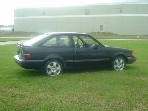 1989 Ford Escort - A black 1989 Ford Escort coupe is parked on a grassy area in front of a large building.