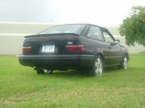 1989 Ford Escort - This black Ford Escort is seen from the rear, parked on a grassy area with a building in the background.