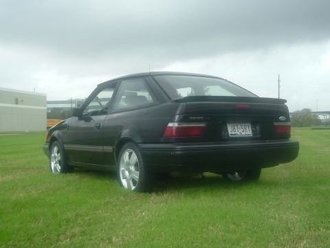 1989 Ford Escort - The rear of a black 1989 Ford Escort coupe is visible, parked on a grassy field under an overcast sky.